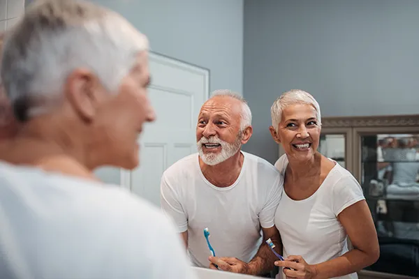 Elderly white couple smiling and brushing their teeth in the bathroom mirror.