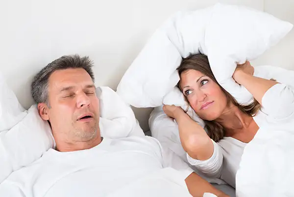 Husband snoring loudly in bed while his wife covers her ears with a pillow due to the noise.